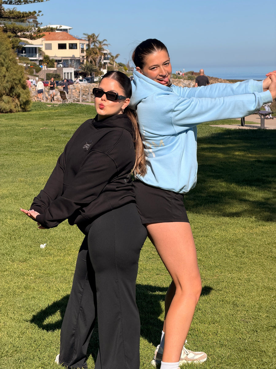 Two friends in black and Alpine Sky CORE O.G. Hoodies on the grass by the beach